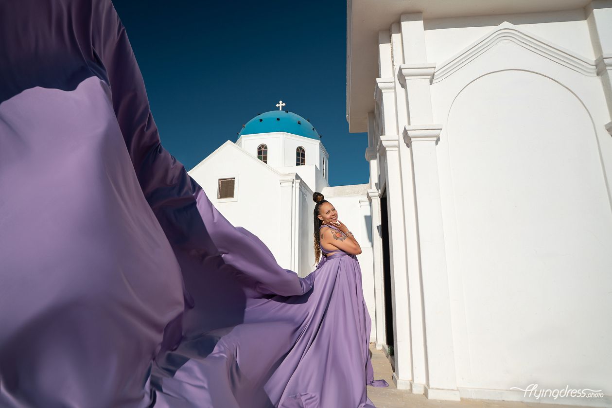 Soft lavender silk fills the frame as a smiling woman poses effortlessly between Santorini's whitewashed walls and iconic blue-domed church.